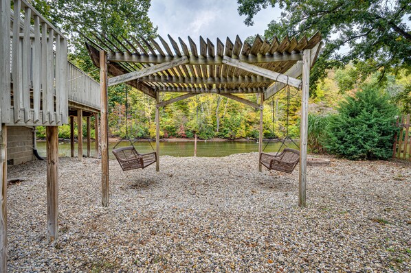 Una pérgola de madera con dos columpios sobre una zona de grava, con vista a un lago y un bosque.