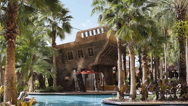 Zona de piscina con palmeras, un edificio con ventanas arqueadas y un paraguas rojo.