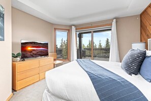 A bedroom with a bed, a TV on a wooden dresser, a balcony with a view of trees, and a window with curtains.