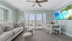 A living room with a sofa, ottomans, a dining table with chairs, a ceiling fan, and a TV on the wall. - Summer Breeze 1-Bed Condo! Gorgeous Views & Pool | Steps to Beach | Book Now! (Miramar Beach)
