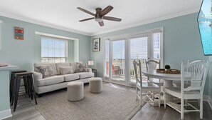 A living room with a sofa, ottomans, a dining table with chairs, a ceiling fan, and a view through the glass doors. - Summer Breeze 1-Bed Condo! Gorgeous Views & Pool | Steps to Beach | Book Now! (Miramar Beach)