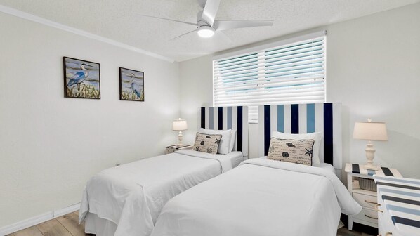 A hotel room with two beds, a ceiling fan, a window with blinds, and artwork on the wall.