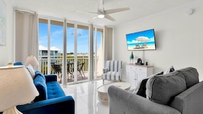 A modern living room with a TV, sofas, a coffee table, and a balcony with a view of buildings and a beach.