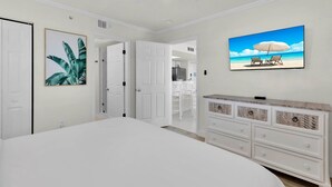 Una camera da letto con un letto grande, una cassettiera con cassetti, un quadro con foglie tropicali e un televisore che mostra una spiaggia.