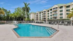 A swimming pool with a white fence, lounge chairs, and a multi-story building with balconies in the background.