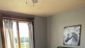 Ein Schlafzimmer mit einem Bett, Vorhängen, einem Gemälde und einem Fenster mit Blick auf Grünfläche.