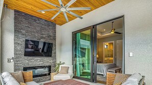 A modern living room with a stone wall, a flat-screen TV, a fireplace, and a sliding glass door leading to a bathroom. - Luxurious 2023 New Construction — Executive Vacation Home Near Vanderbilt Beach (Naples)