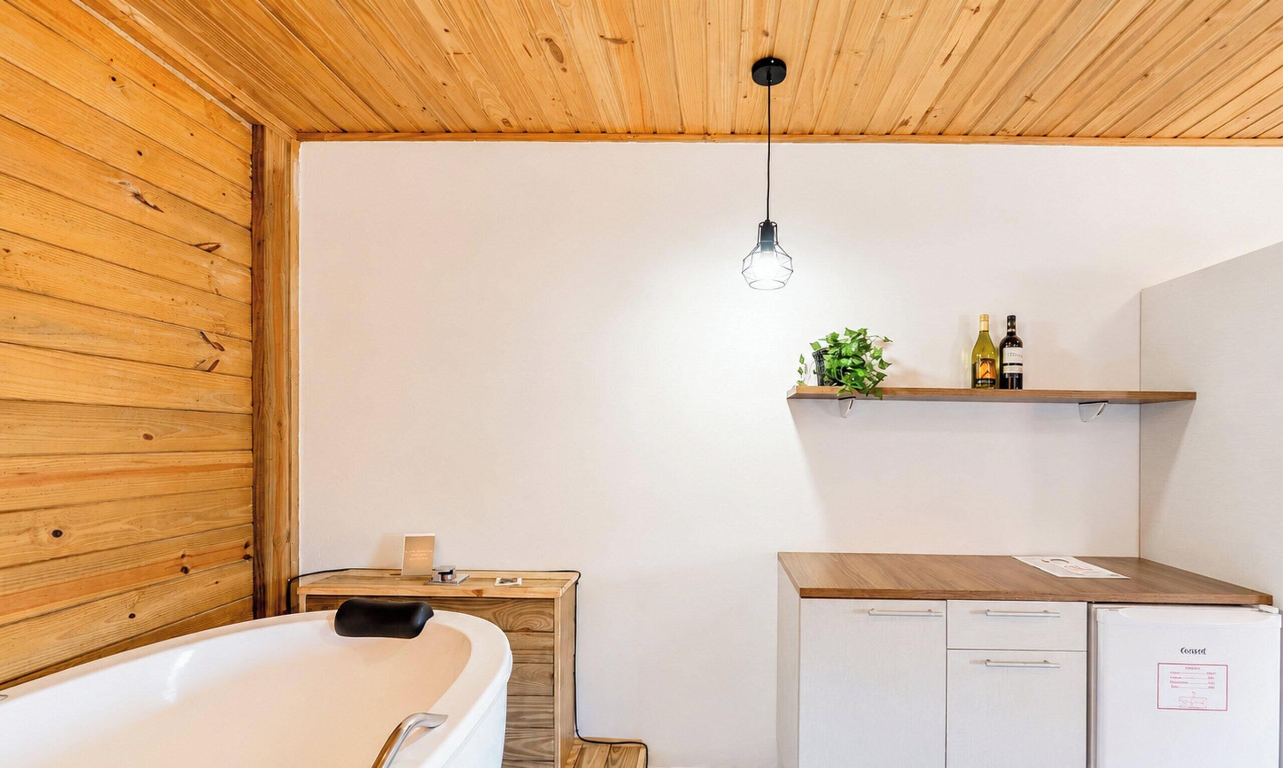 A modern bathroom with a wooden ceiling, a bathtub, a countertop with a sink, and a small refrigerator.