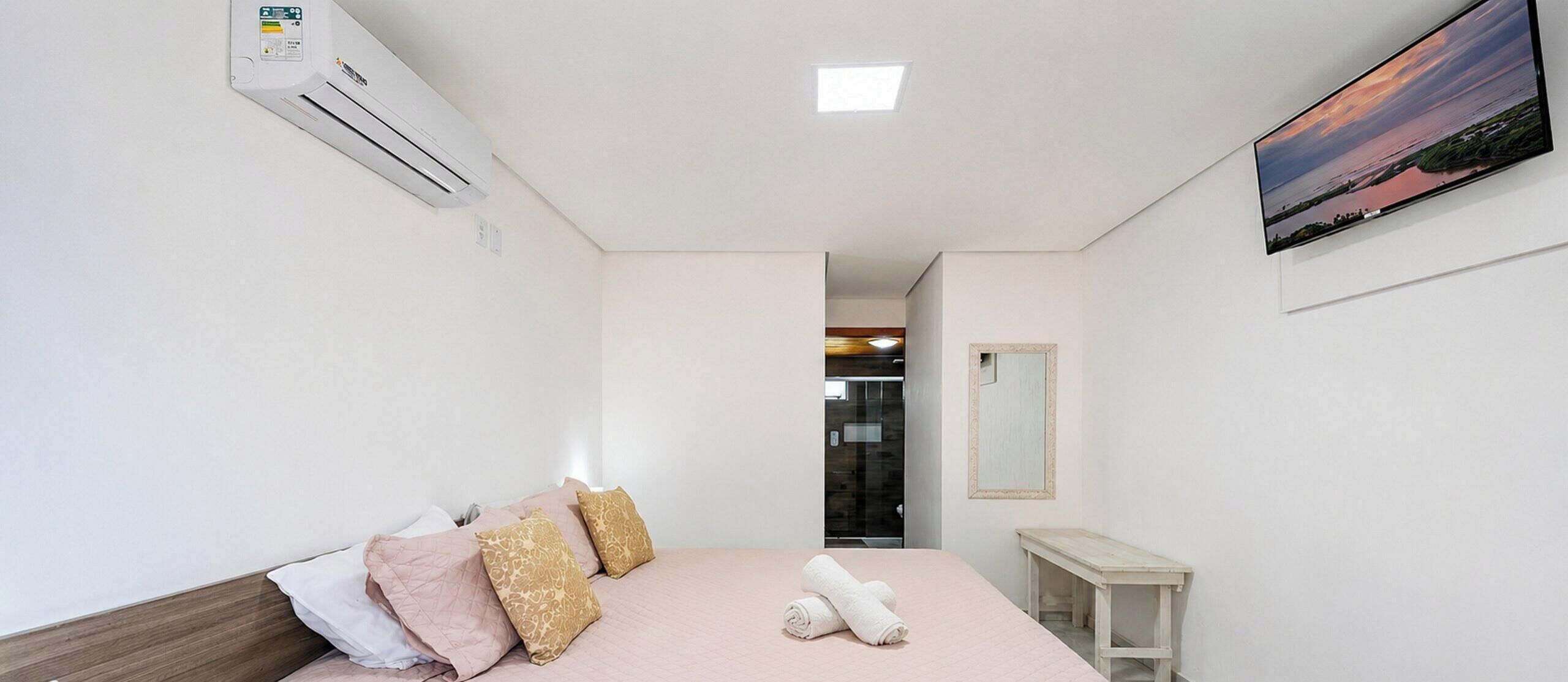 A hotel room with a bed, pillows, an air conditioning unit, a painting, and a mirror.