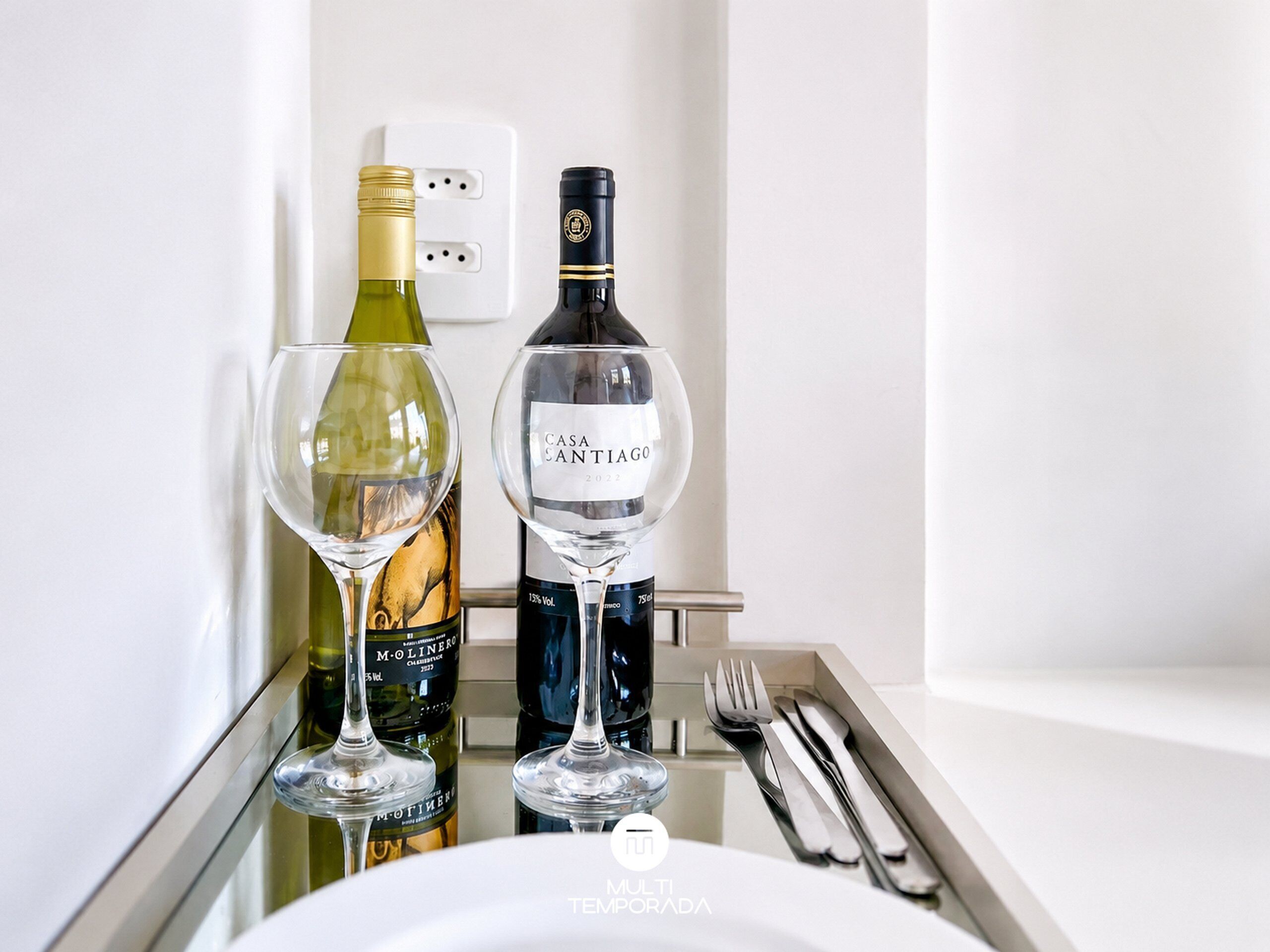 A minibar with two wine bottles and two glasses, a power outlet, and a set of cutlery on a tray.