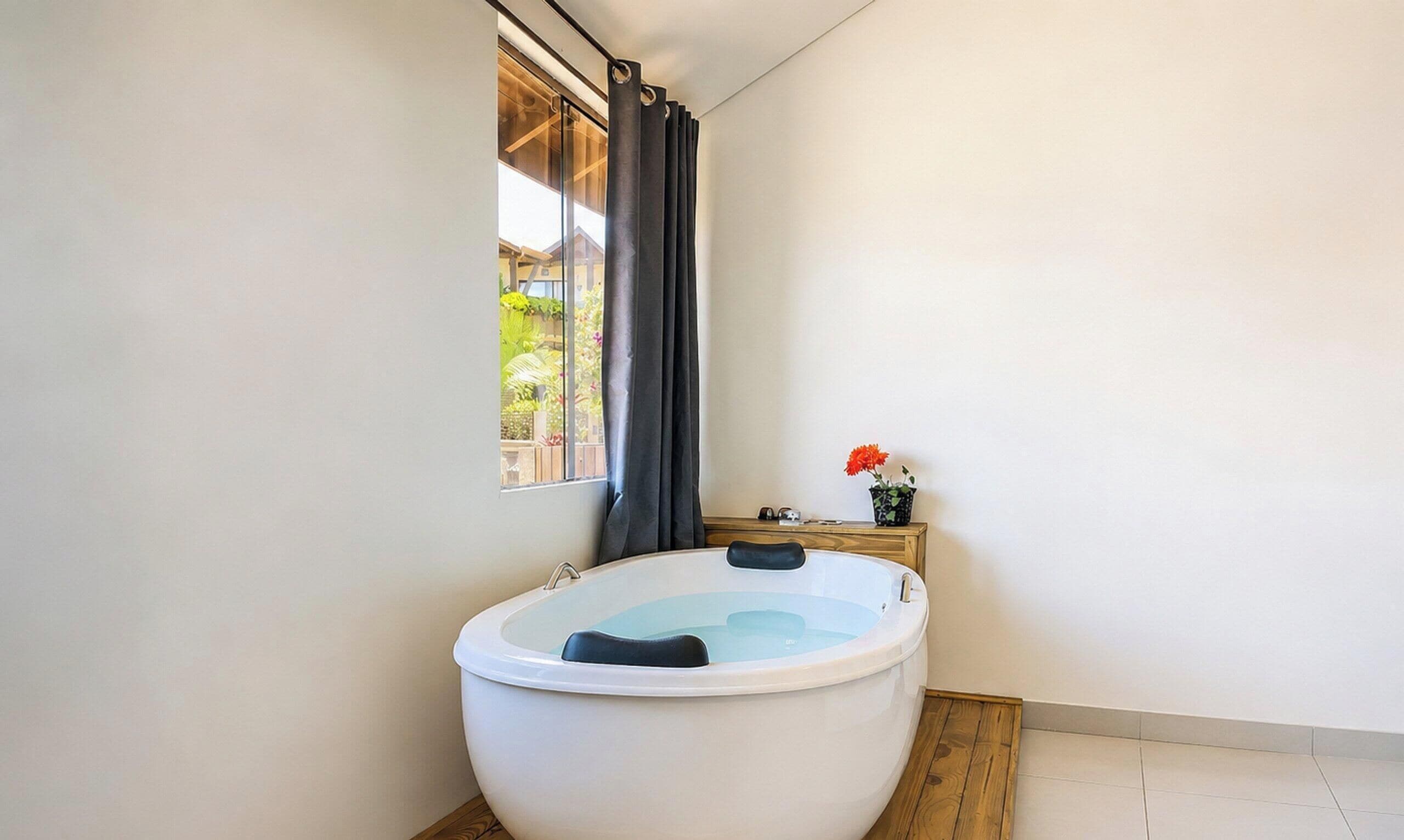A bathroom with a large bathtub, a window with curtains, and a small wooden side table with a flower vase.