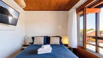 Quarto com cama, almofadas, mesa de cabeceira, televisão e janela com cortinas.