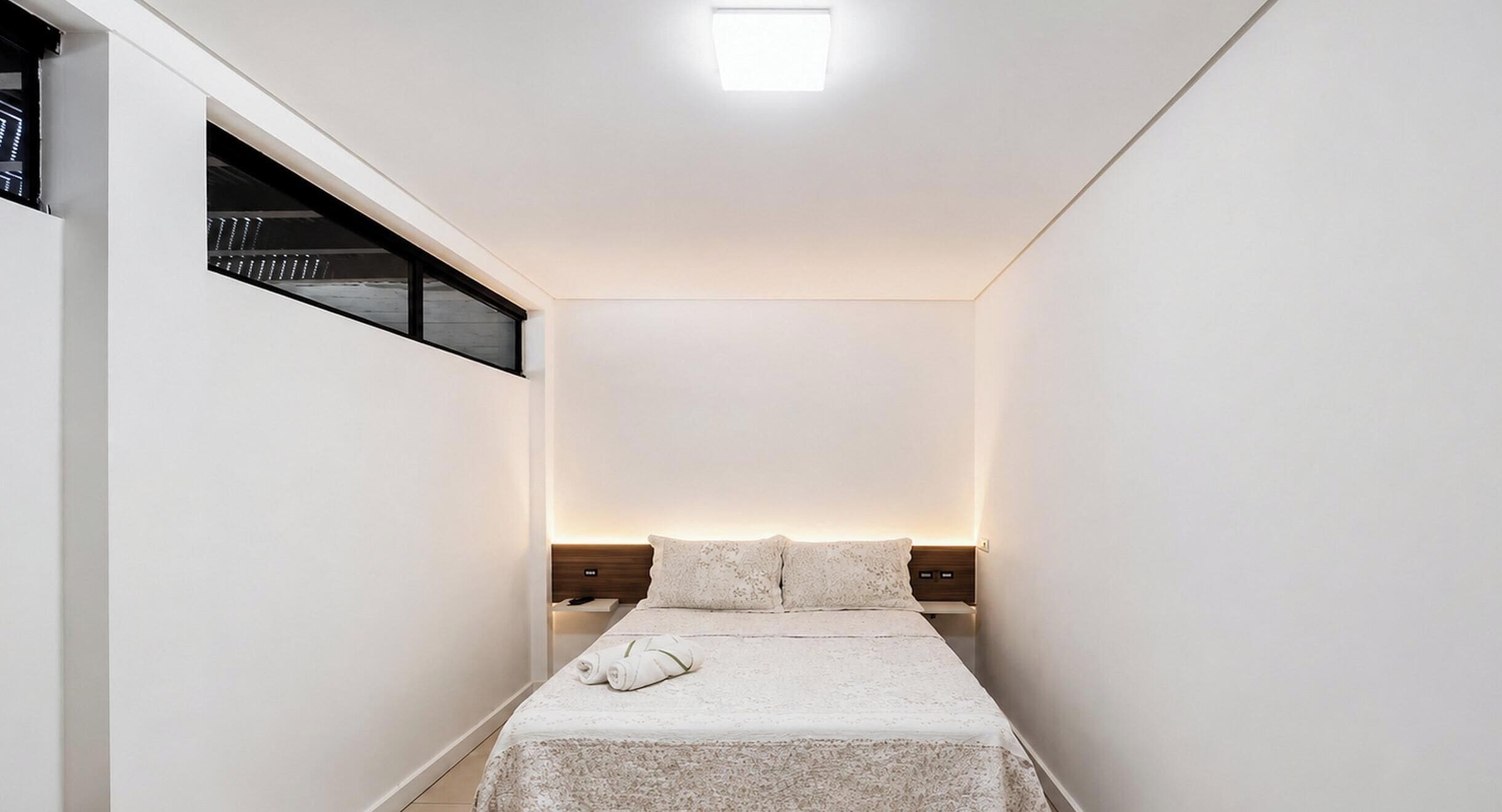 A narrow hotel room with a bed, a nightstand, a wall-mounted shelf, and a ceiling light.