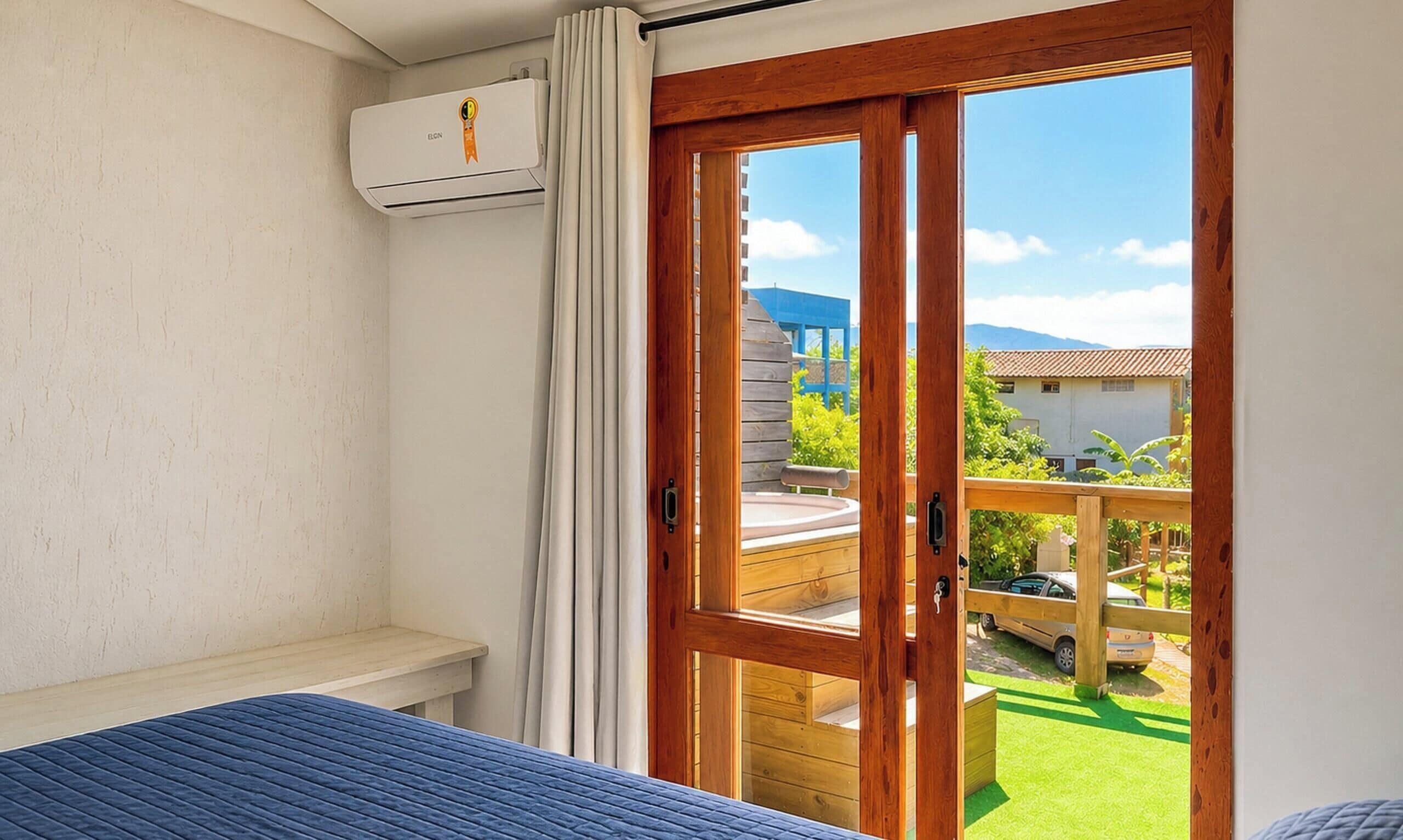 Un dormitorio con cama, equipo de aire acondicionado, una puerta de madera que da a un balcón y vistas al exterior.