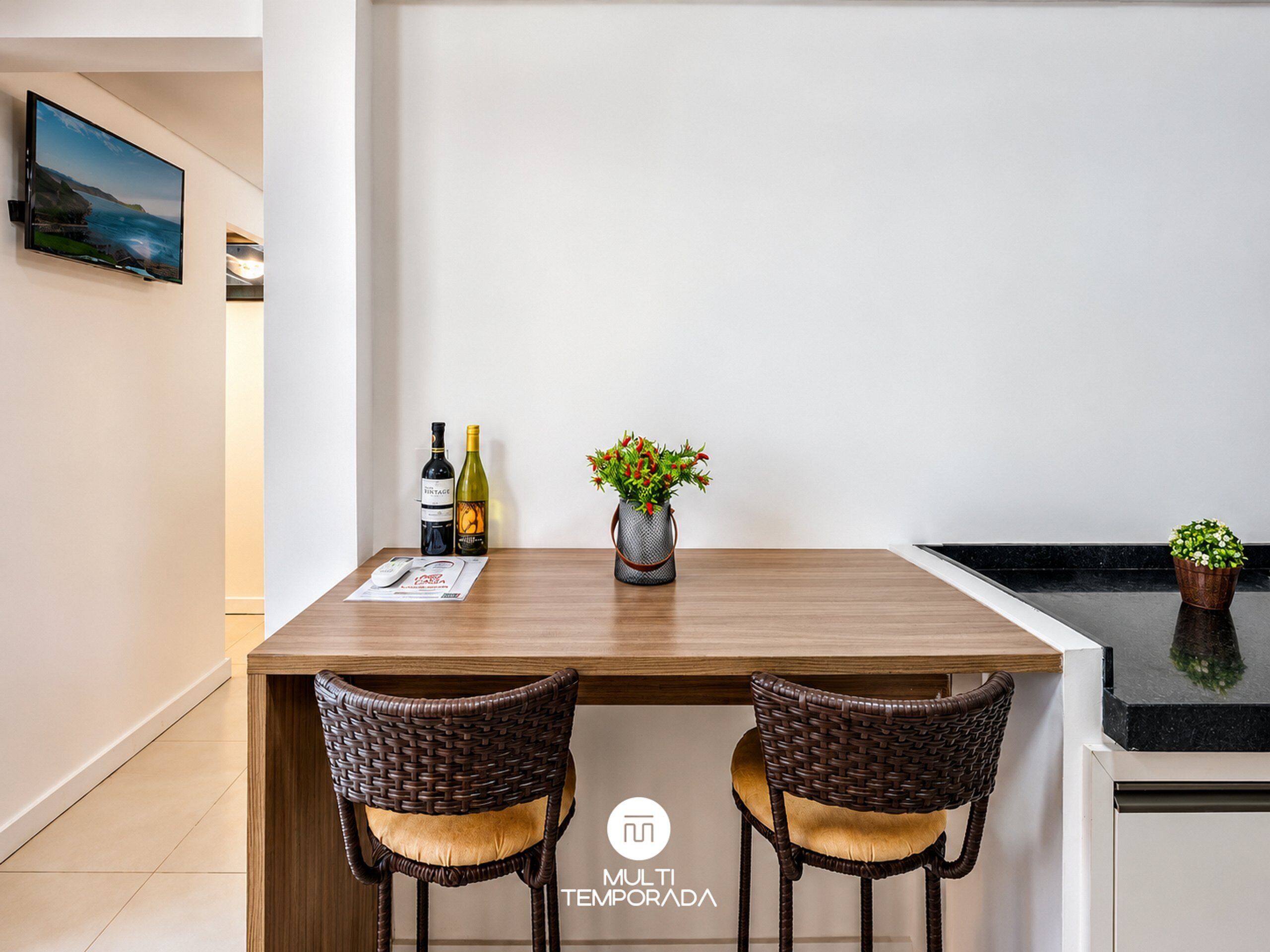 A modern dining area with a wooden table, two wicker chairs, a wine bottle, a potted plant, and a TV mounted on the wall.