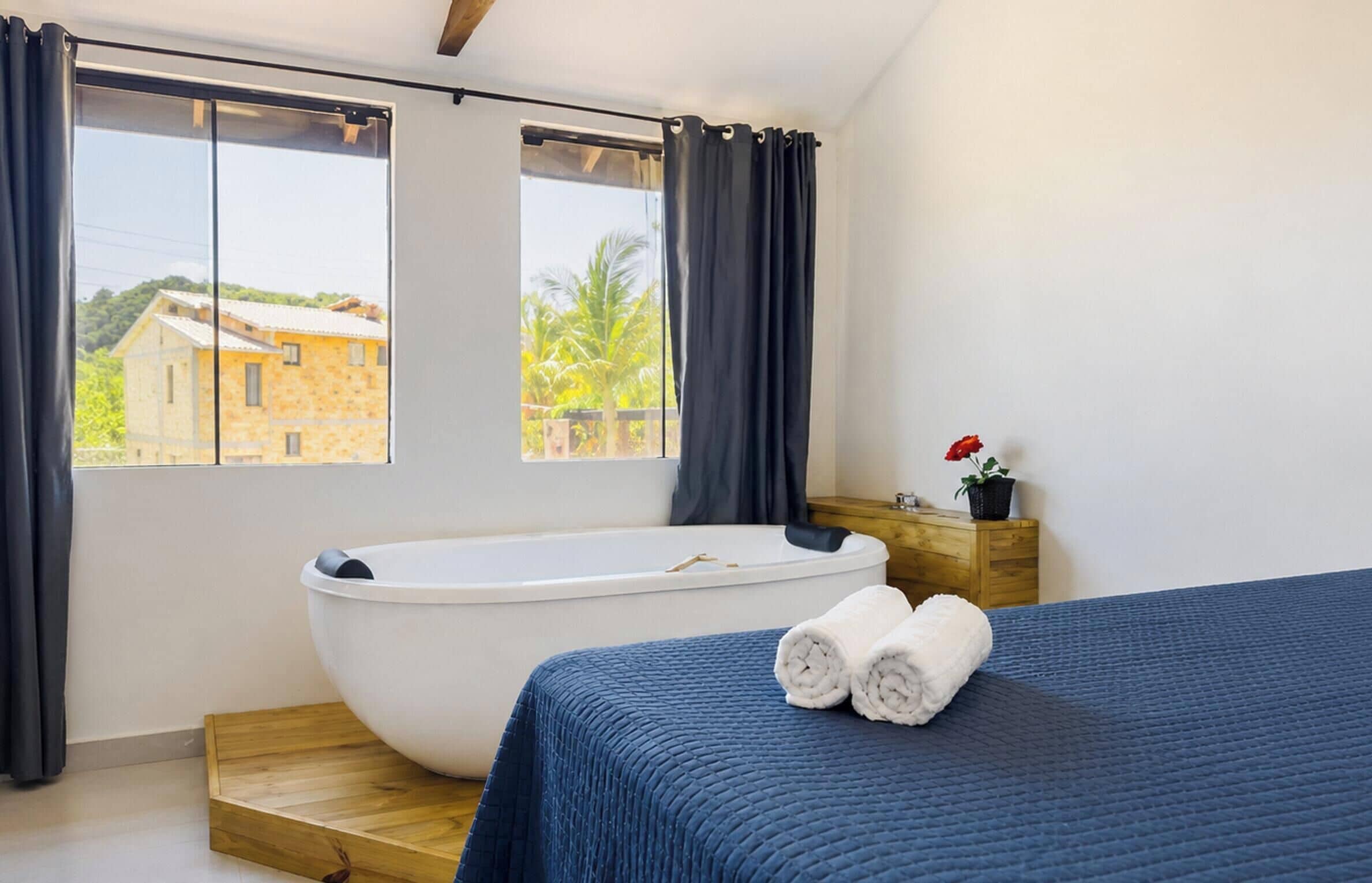 A bedroom with a large bathtub, a bed with a blue bedspread, and a view of a building outside.