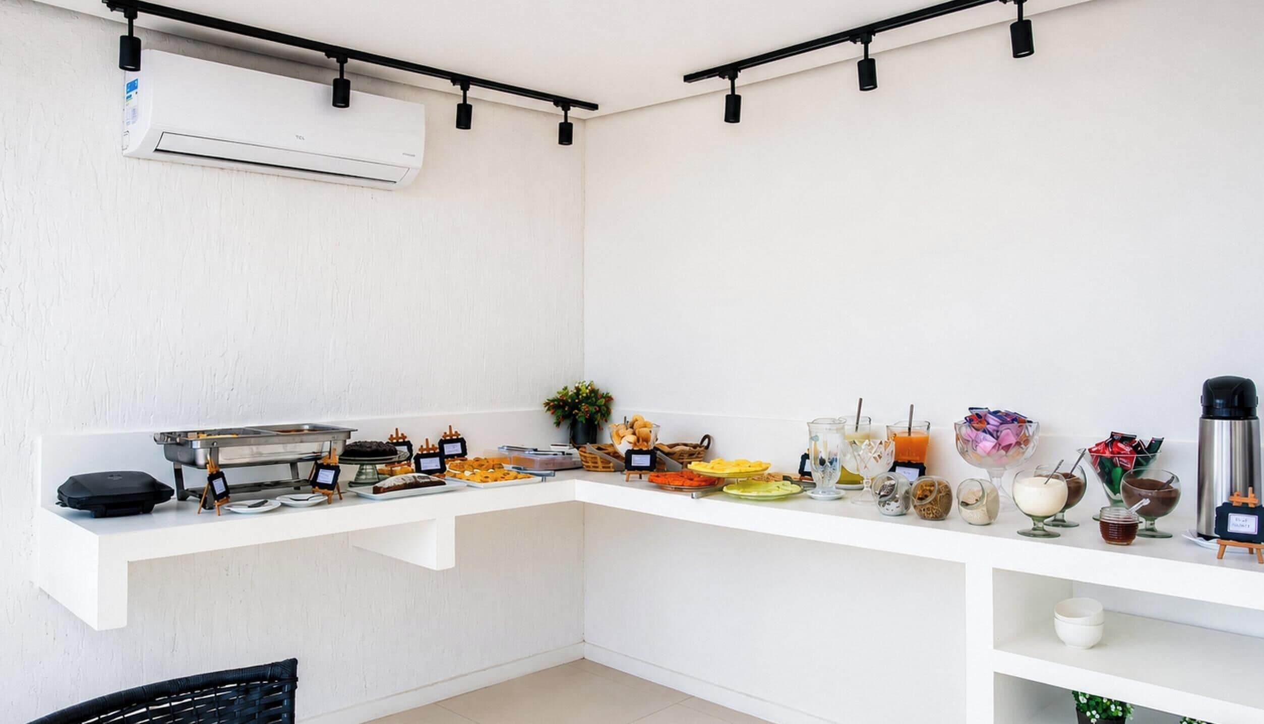 A kitchenette with a built-in air conditioner, a coffee machine, and a variety of food and drink options on a white countertop.