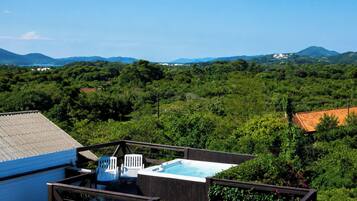 A hot tub on a deck with chairs, surrounded by lush greenery and mountains in the background.
