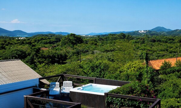 A hot tub on a deck with chairs, surrounded by lush greenery and mountains in the background.