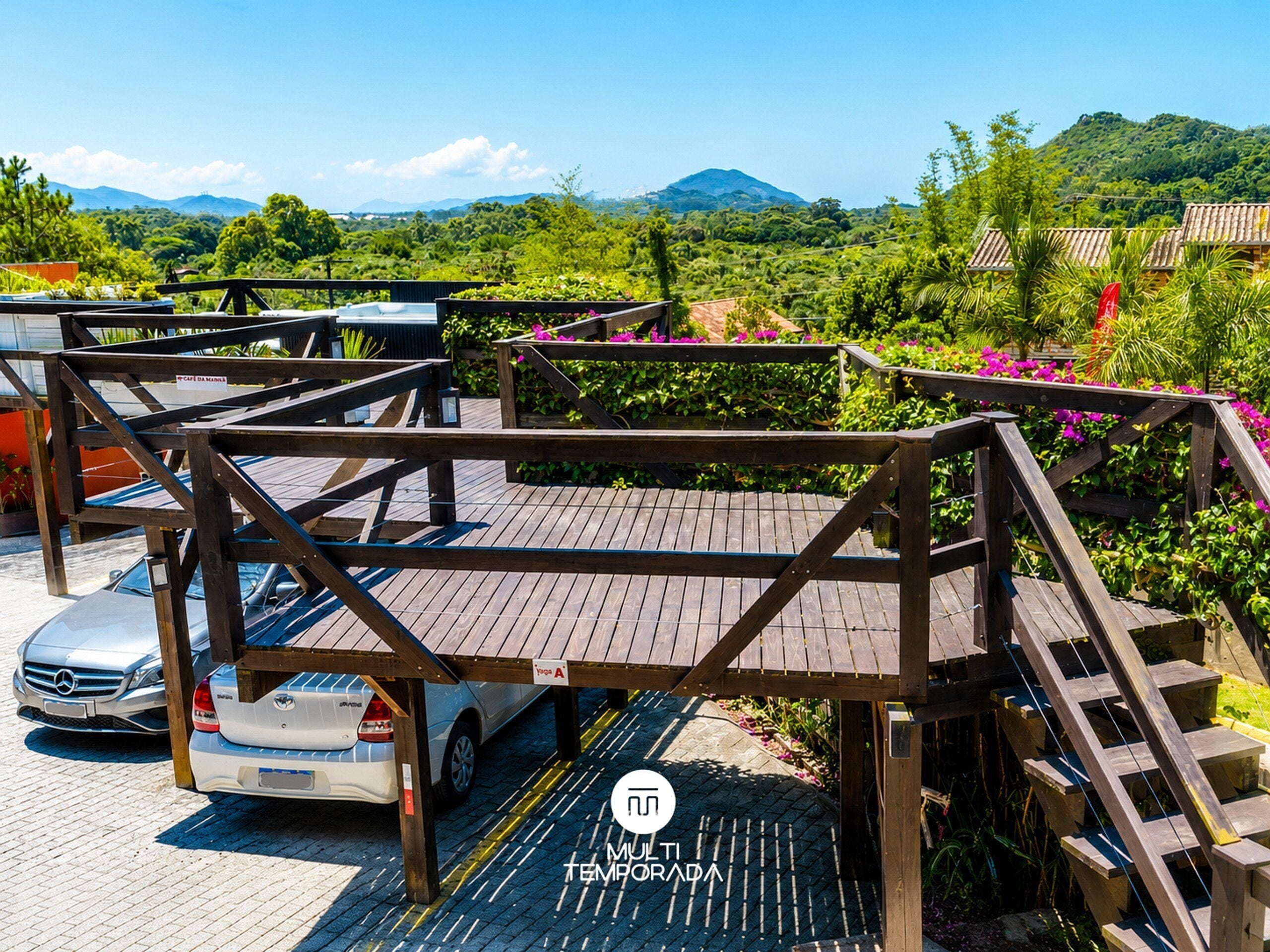 A wooden deck with a car parked underneath, surrounded by lush greenery and a mountainous backdrop.