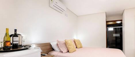 Habitación de hotel con cama, almohadas, mesita de noche con una botella de vino y copas, y aire acondicionado instalado en la pared.