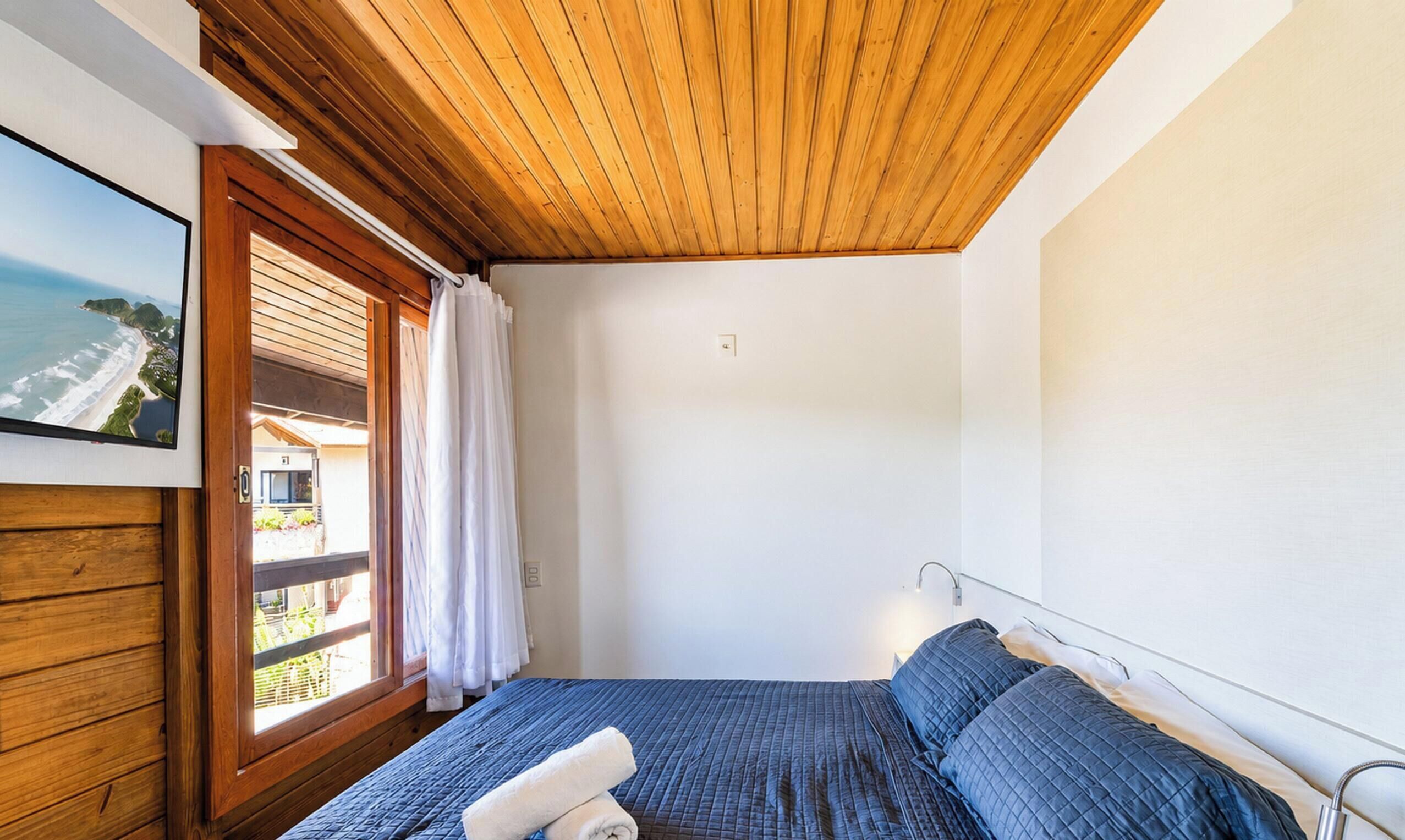 Un dormitorio con techo de madera, cama con ropa de cama azul, ventana con cortinas y vista a la playa.