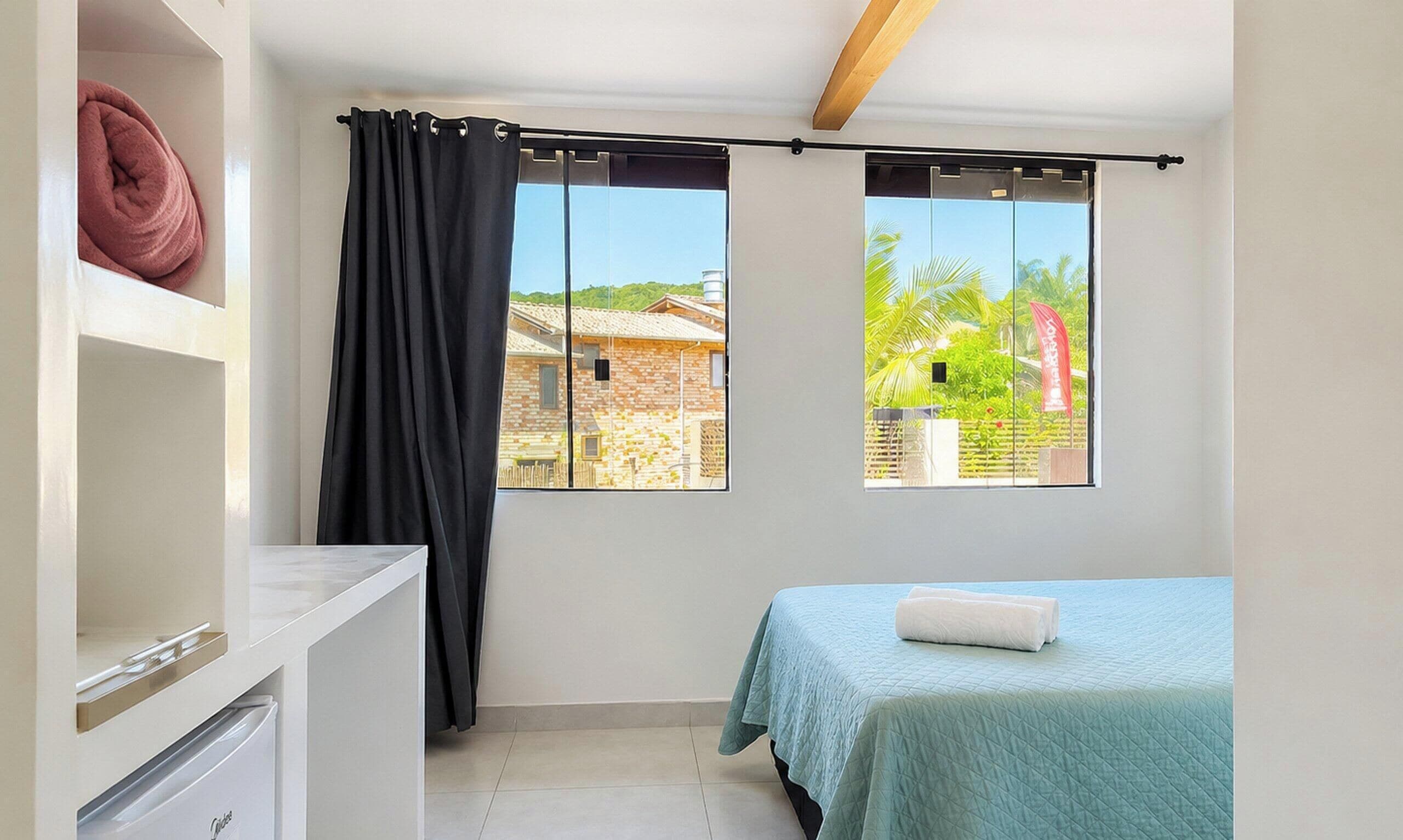 Une chambre à coucher avec un lit, un petit bureau, une fenêtre avec des rideaux et une vue sur un immeuble et de la verdure à l’extérieur.