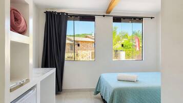 Un dormitorio con cama, un escritorio pequeño, una ventana con cortinas y vistas a un edificio y vegetación.