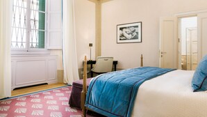 Een slaapkamer met een bed, een raam met luiken, een lijst met een afbeelding aan de muur, en een kamerbreed tapijt met dessin.