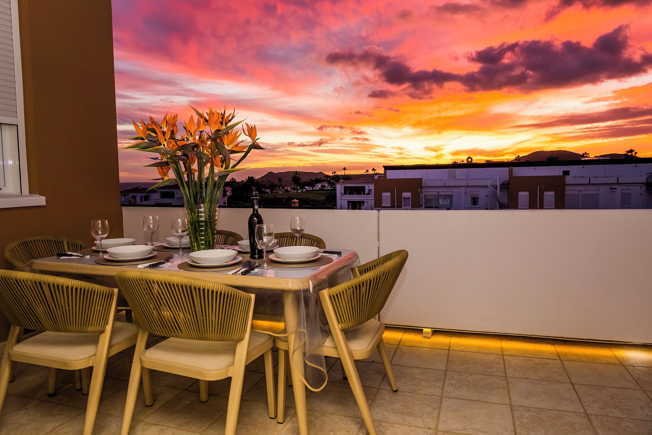 Une table à manger dressée pour quatre personnes, avec des chaises, un vase de fleurs et une vue sur un coucher de soleil au-dessus d’un quartier résidentiel.
