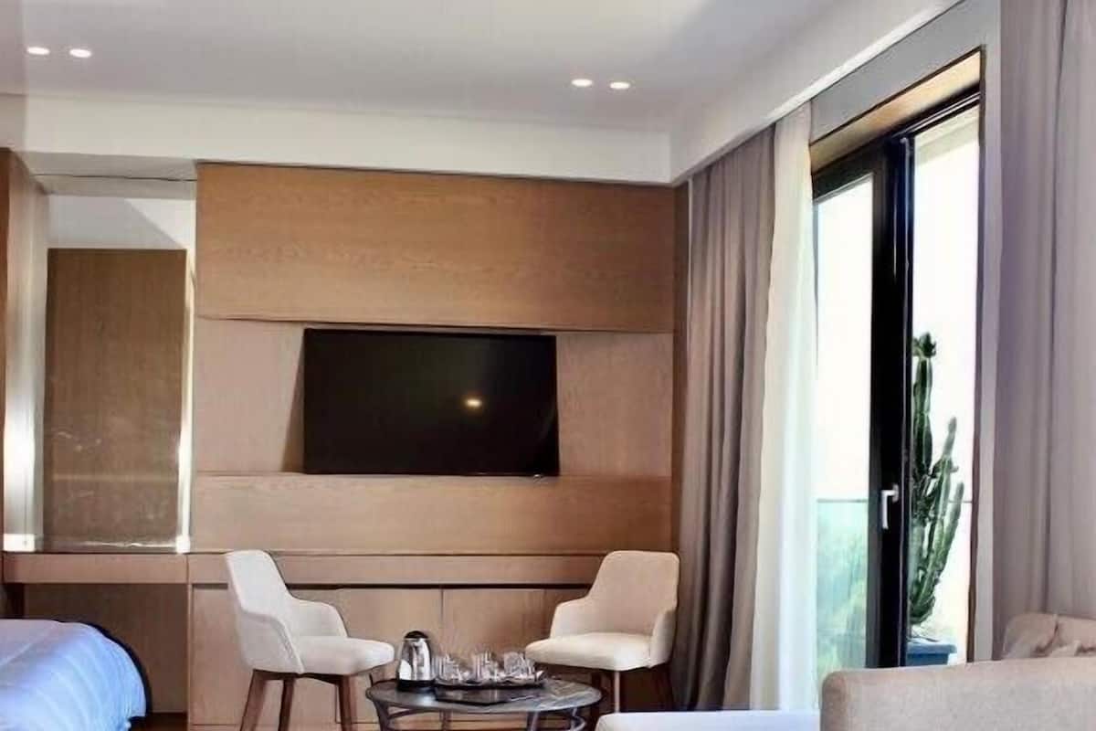 Hotelkamer met een groot bed, twee fauteuils, een klein rond tafeltje, een flatscreen-tv aan de muur en een groot raam met gordijnen.
