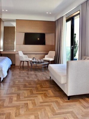 A hotel room with a large bed, two armchairs, a small round table, a flat-screen TV mounted on the wall, and a large window with curtains.