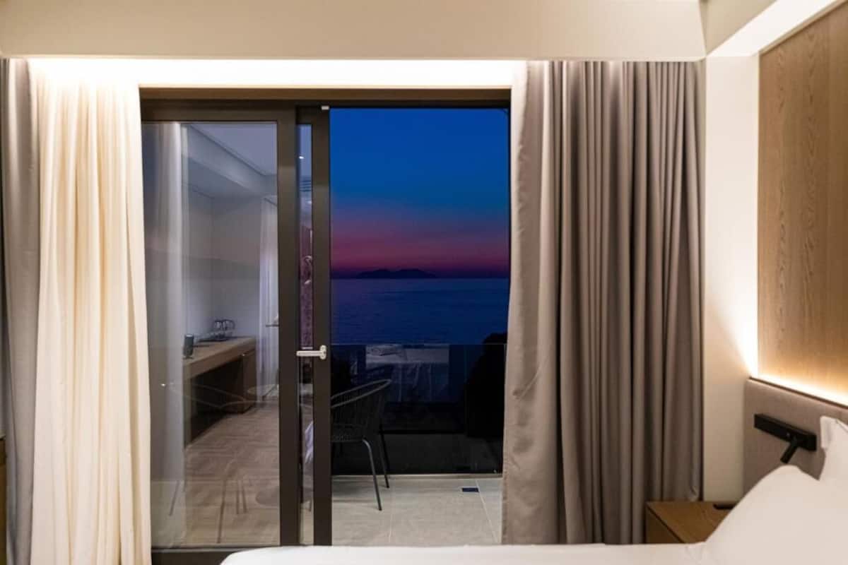 Een hotelkamer met een bed, een bureau en een stoel. Door een glazen deur is er uitzicht op de oceaan.