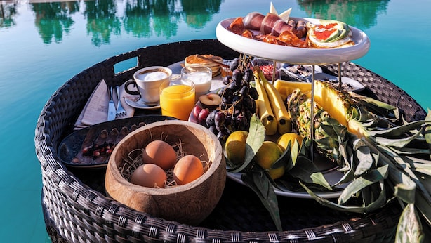 Un plateau de petit-déjeuner avec des œufs, des fruits et des viennoiseries, placé au bord de la piscine, offrant une vue dégagée sur l’océan et les arbres.