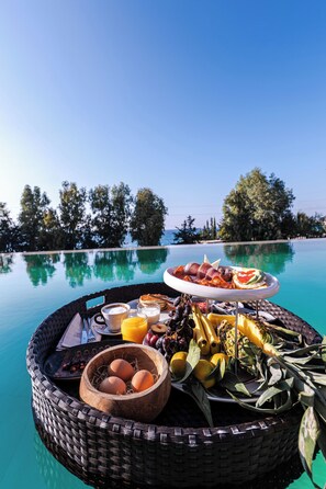 Ein Frühstücksbrett mit Eiern, Früchten und Gebäck am Pool mit freiem Blick auf das Meer und Bäume.