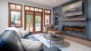 A modern living room with a fireplace, leather sofa, and a large window overlooking a forested area.