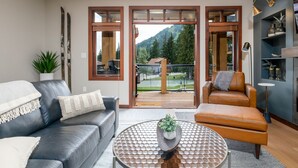 A modern living room with a sofa, armchair, and coffee table, featuring a view of a tennis court and mountains.