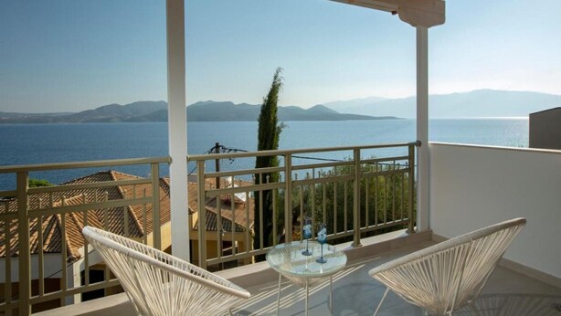 Un balcón con vistas despejadas al mar y las montañas, muebles de mimbre blanco y una mesa de vidrio.