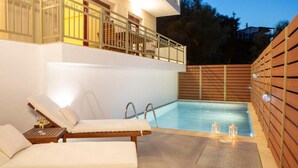 Un área de piscina moderna al aire libre con dos sillones de descanso y una mesita, rodeada de cerramiento de madera y un balcón con barandillas.