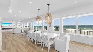 Un vaste salon doté d’une grande table à manger blanche, de chaises blanches et d’un téléviseur fixé au mur. De grandes fenêtres offrent une vue sur l’océan.
