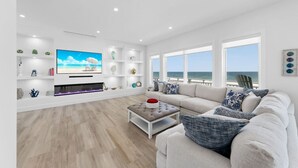 Un salon moderne doté d’un téléviseur à écran plat, d’étagères blanches, d’un canapé beige et offrant une vue sur l’océan.
