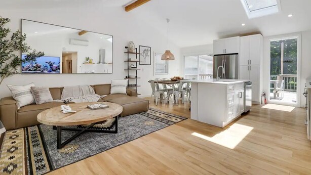 A spacious living area with a sofa, coffee table, and a dining table. There is a kitchen with white cabinets and a small island. A large mirror and a hanging light fixture are visible. - Modern Rustic Private Ranch w/ Peloton! (Henryville)