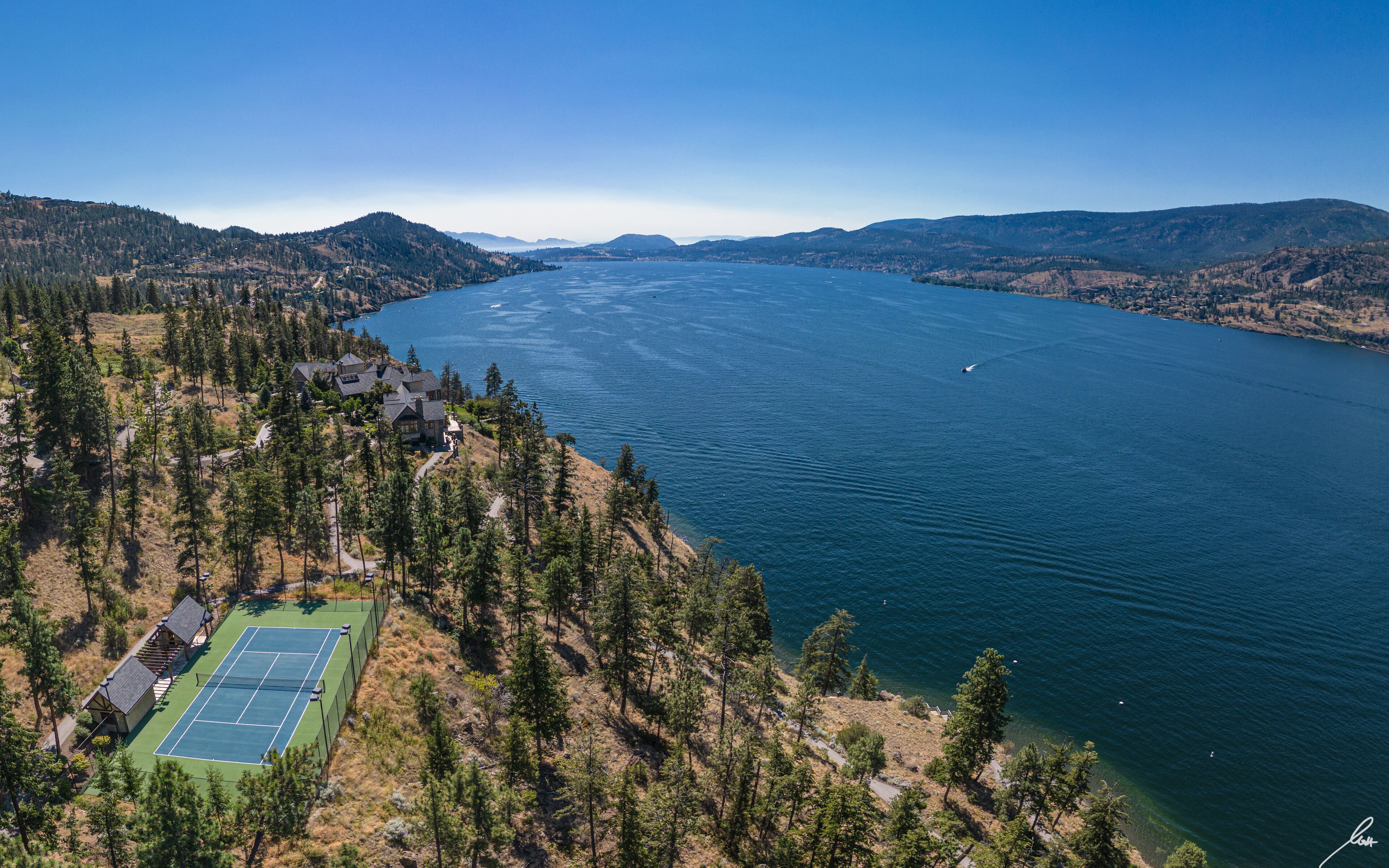 An aerial view of a lakeside property with a tennis court, surrounded by dense forest and hills.