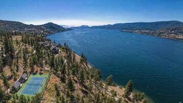An aerial view of a lakeside property with a tennis court, surrounded by dense forest and hills.
