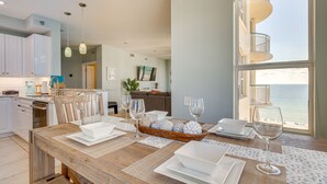 A modern kitchen with white cabinets, a dining area with a wooden table set for four, and a view of the ocean through large windows.