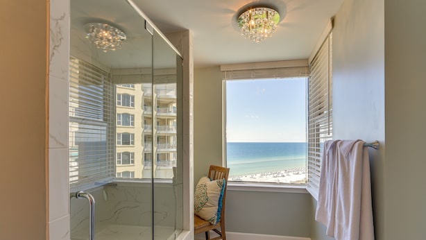A bathroom with a glass shower, a view of the beach, and a chair with a cushion.