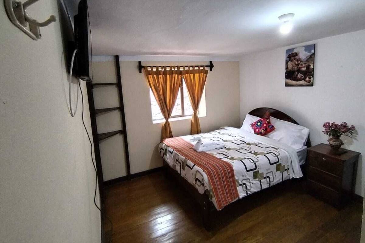 A bedroom with a bed, a nightstand, a wall-mounted TV, a window with curtains, and a ceiling light.