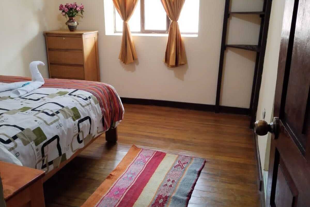 A bedroom with a bed, a TV mounted on the wall, a wooden dresser, and a window with curtains.