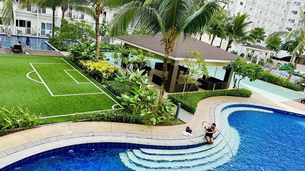 Een zwembad met een golvend ontwerp, een kleine tuin met palmbomen en een voetbalveld.