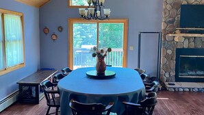 Un coin repas avec une table ronde, des chaises et un lustre. On y trouve également une cheminée en pierre et une fenêtre donnant sur un paysage verdoyant.